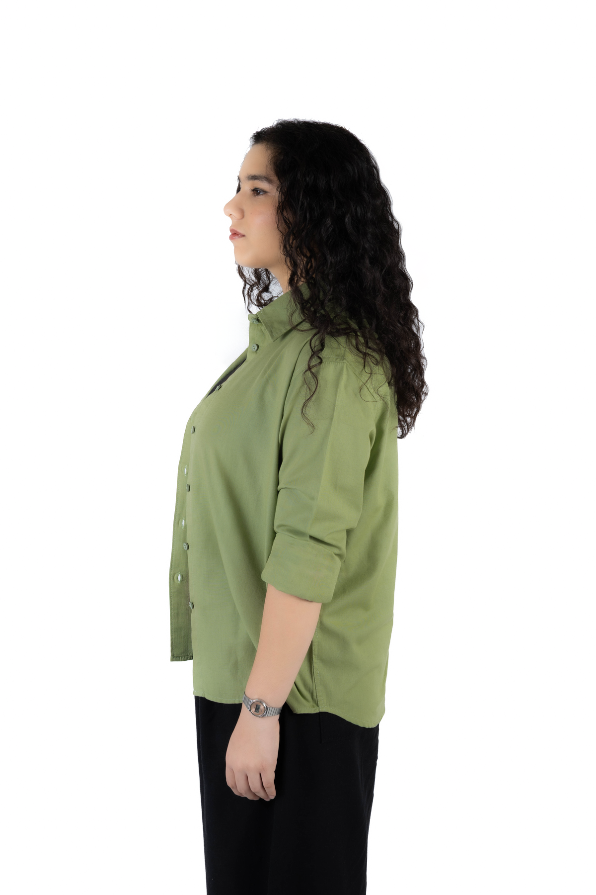 A woman with long, curly dark hair is shown in profile wearing a sage green button-up shirt with rolled-up sleeves and black pants, photographed against a solid white background.