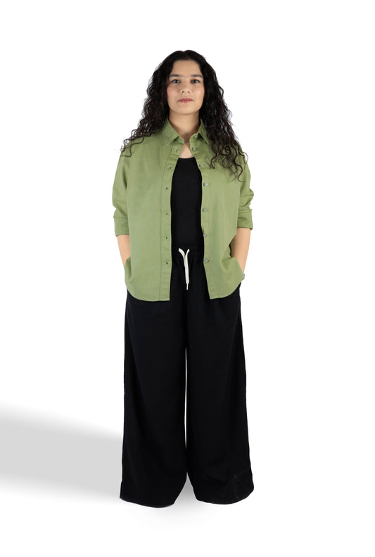 A woman stands against a white studio background wearing a light olive green button-up shirt over a black top and black wide-leg pants.