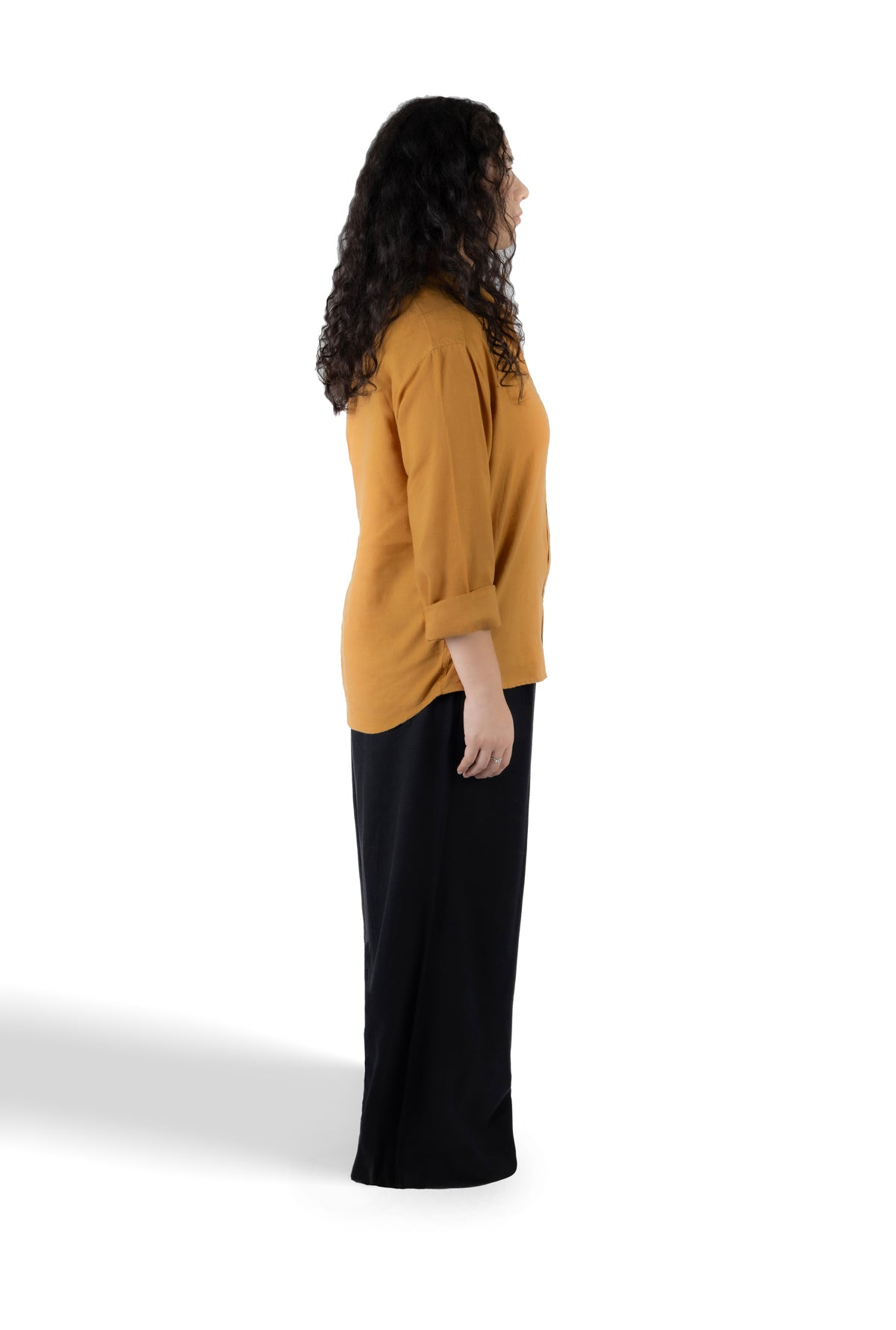 A woman shown in side profile wearing a mustard yellow long-sleeve top and black pants against a plain white studio background.