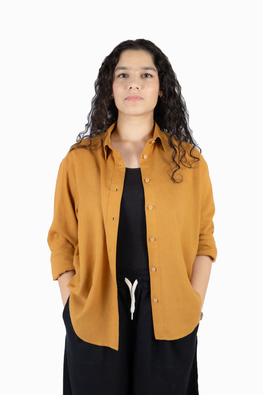 A woman with long, curly dark hair wearing a mustard yellow linen button-up shirt over a black top and black pants with a white drawstring, standing against a solid white background.