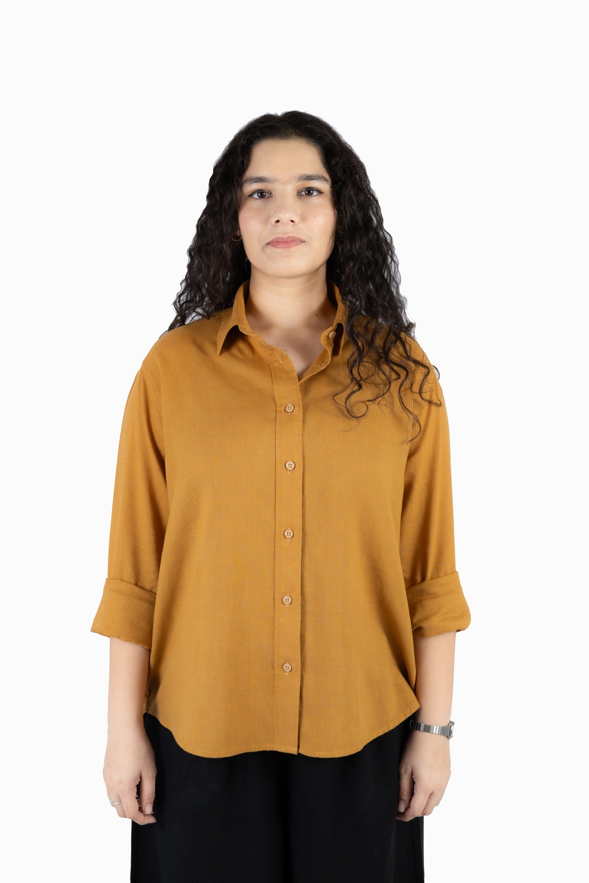 A woman with long, curly dark hair wearing a mustard yellow button-up shirt with rolled-up sleeves and black pants, standing against a plain white background.