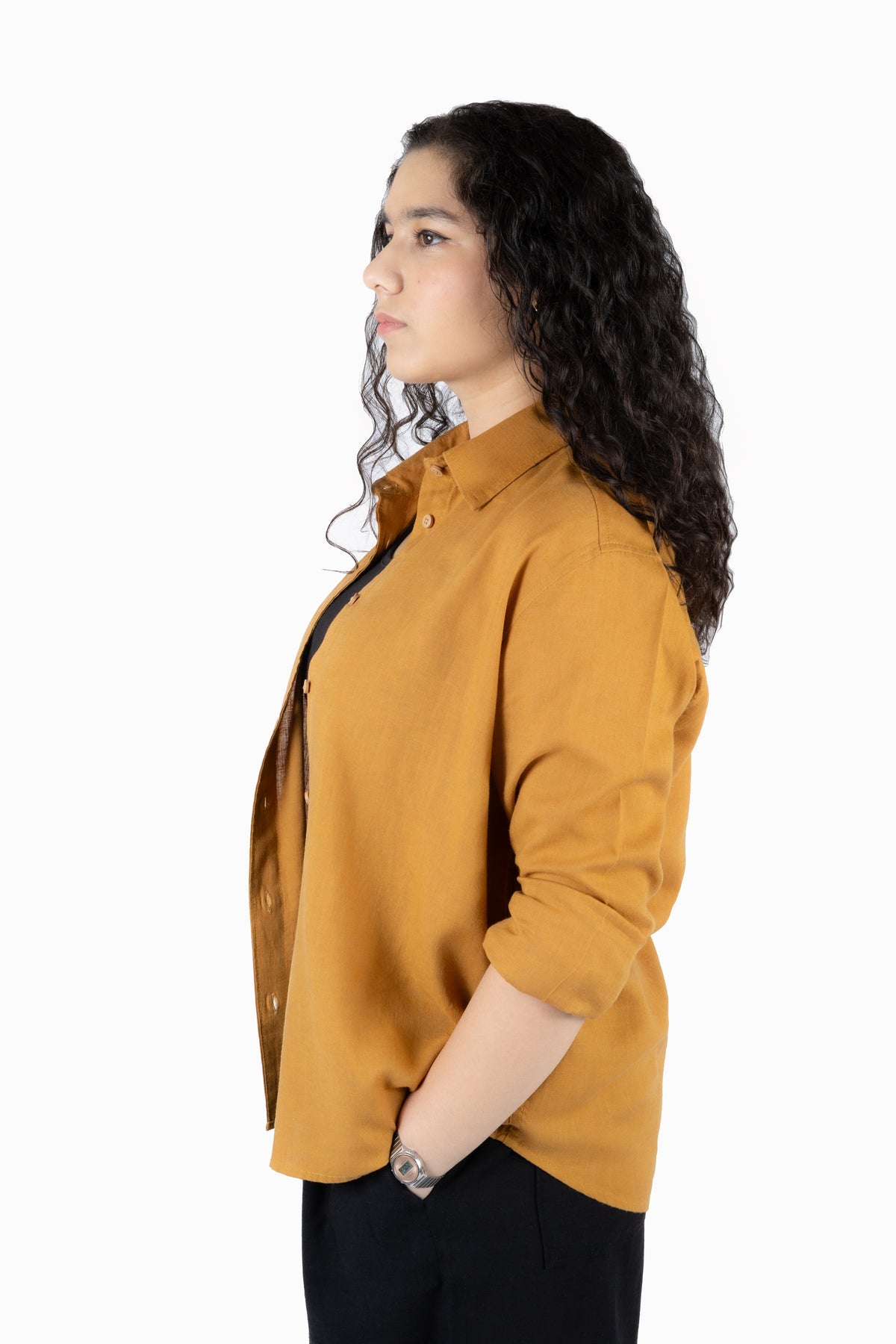 A woman with long, curly dark hair is shown in profile wearing a mustard yellow button-down shirt and black pants against a plain white background.