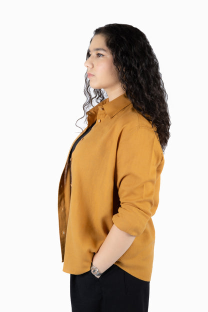 A woman with long, curly dark hair is shown in profile wearing a mustard yellow button-down shirt and black pants against a plain white background.