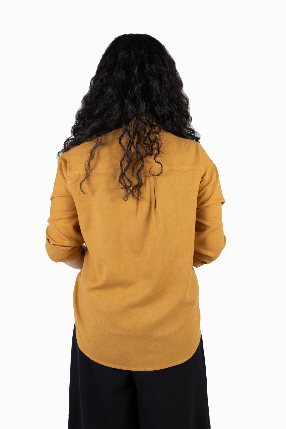 A woman with long, curly black hair is shown from behind wearing a mustard yellow long-sleeve top and black pants against a plain white background.