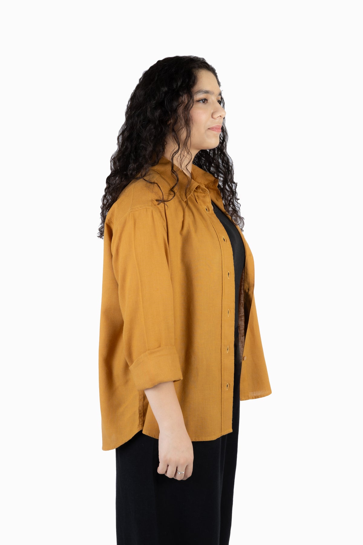A woman with long, curly dark hair is shown in profile wearing a mustard yellow button-up shirt over a black dress, photographed against a solid white studio background.