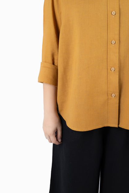 A close-up of a person wearing a mustard yellow button-front shirt with rolled sleeves and black pants, shown against a plain white background.