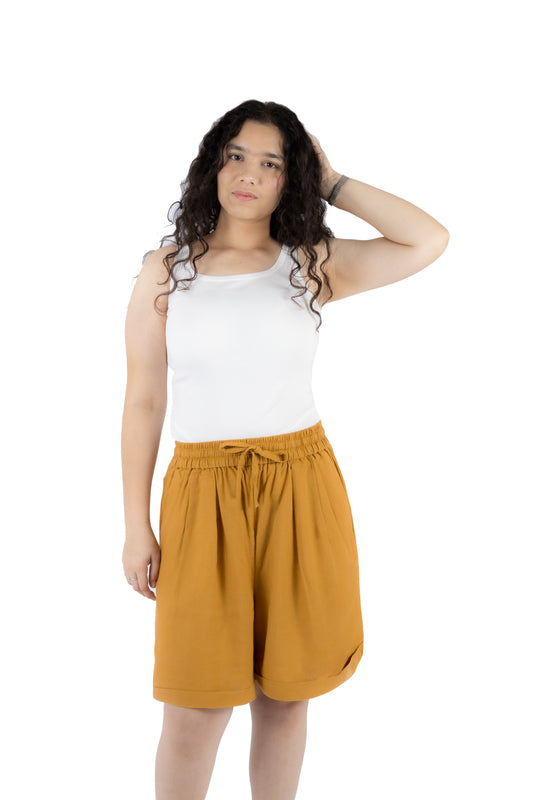 A woman modeling a pair of mustard yellow drawstring shorts and a white tank top against a solid white background.