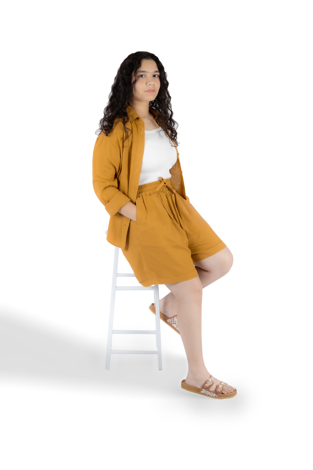 A woman with long curly dark hair sits on a white stool against a white studio background, wearing a mustard yellow linen-blend outfit consisting of a button-up shirt, matching shorts, and a white tank top, paired with tan slide sandals.