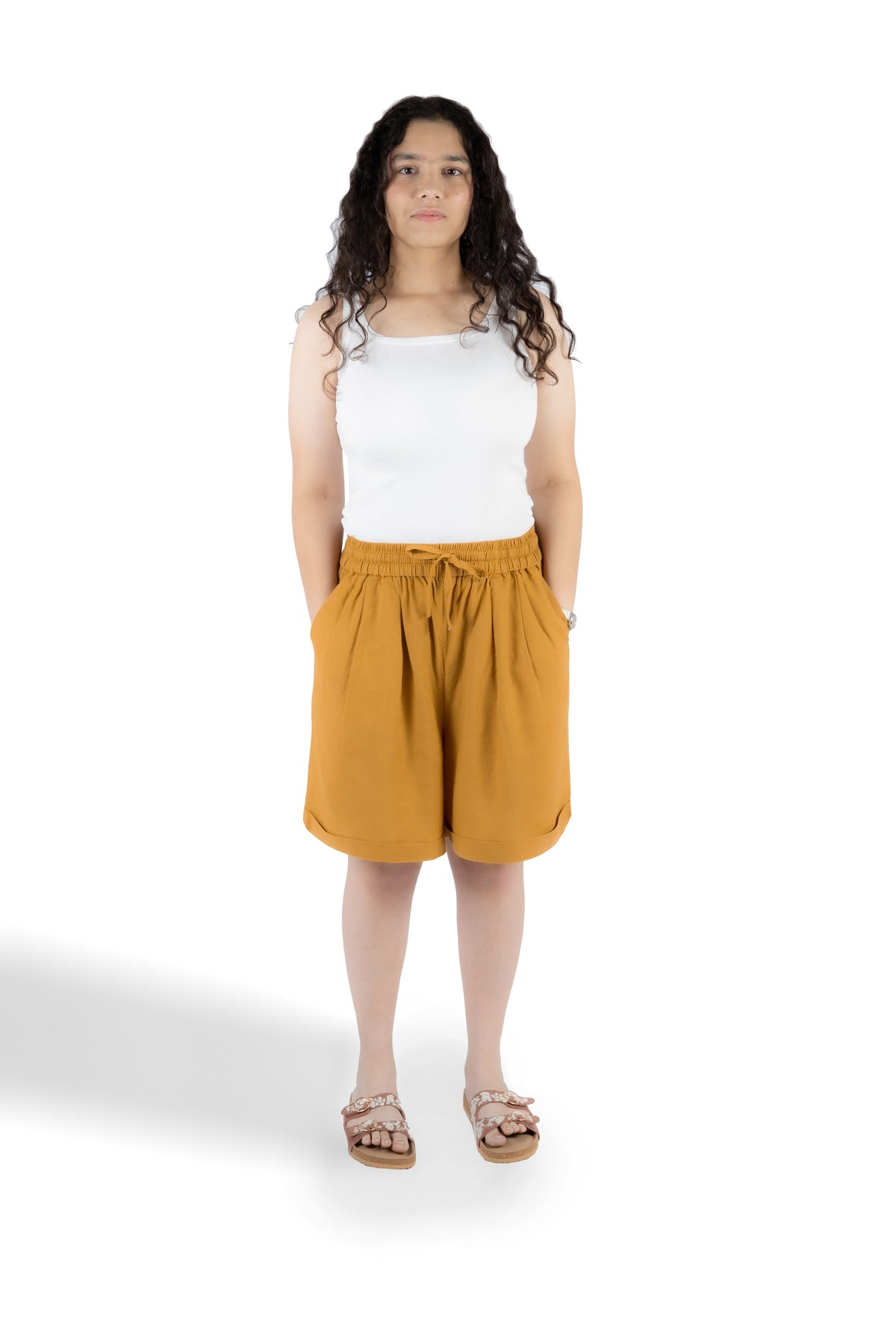 A woman modeling a pair of mustard yellow drawstring shorts with a white tank top and sandals, standing against a solid white background.