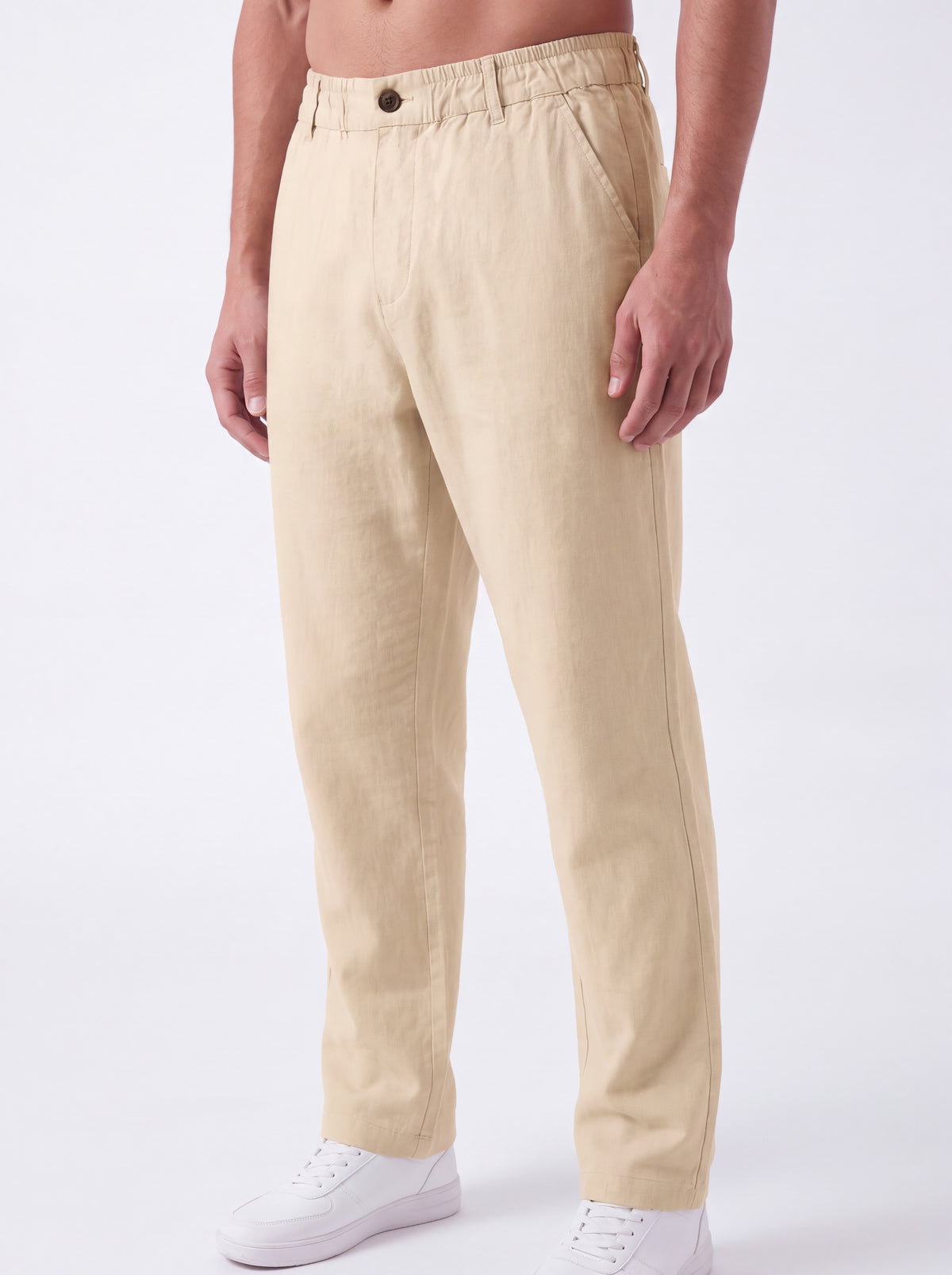 A man wearing beige linen trousers and white sneakers, shown from the waist down against a plain white studio background.