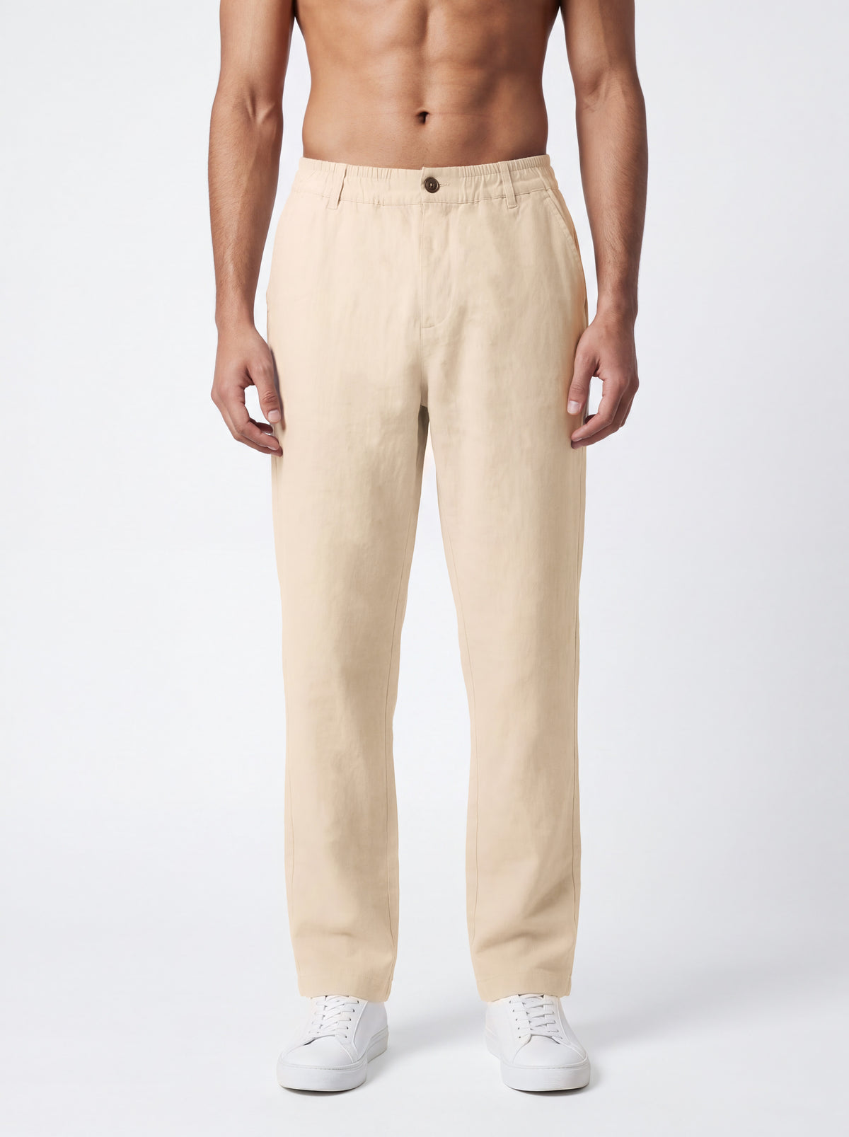 A man wearing beige straight-leg trousers and white sneakers, shown from the waist down against a light gray studio background.