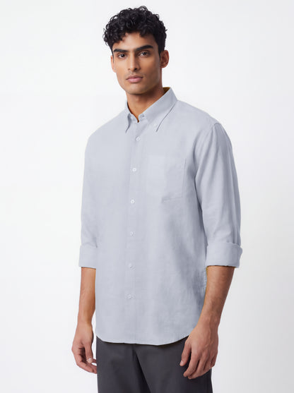 A man wearing a light gray long-sleeve button-down shirt with rolled-up sleeves and dark gray trousers, standing against a plain white studio background.
