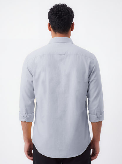 A man viewed from the back wearing a light gray long-sleeve button-down shirt with rolled-up sleeves and black pants, set against a plain white studio background.