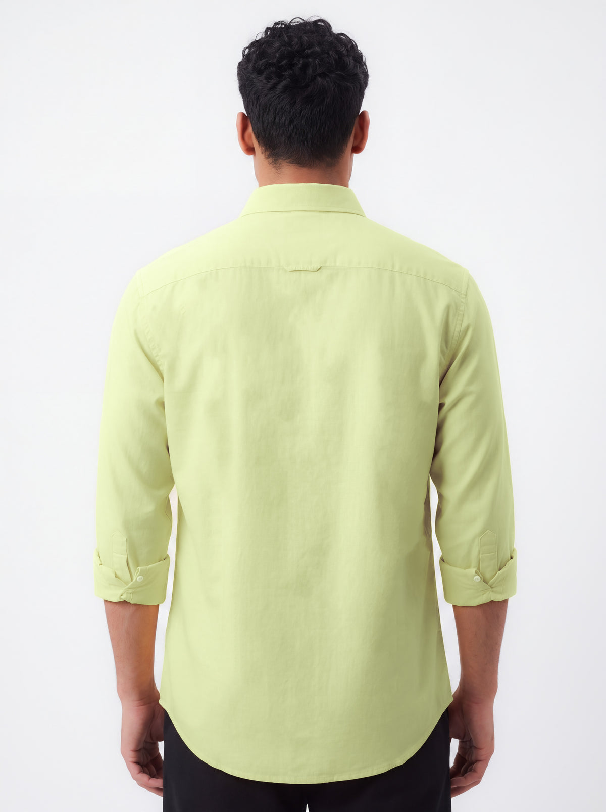 A man viewed from the back wearing a light yellow-green long-sleeve button-up shirt with rolled-up sleeves and black pants, set against a plain white background.