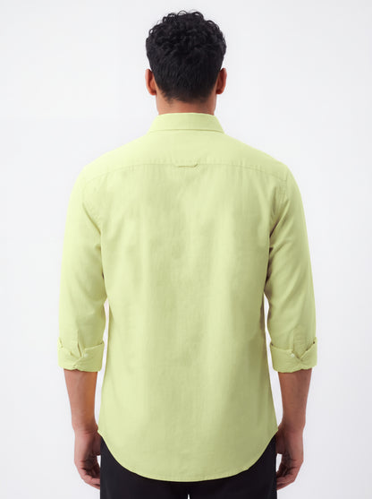 A man viewed from the back wearing a light yellow-green long-sleeve button-up shirt with rolled-up sleeves and black pants, set against a plain white background.