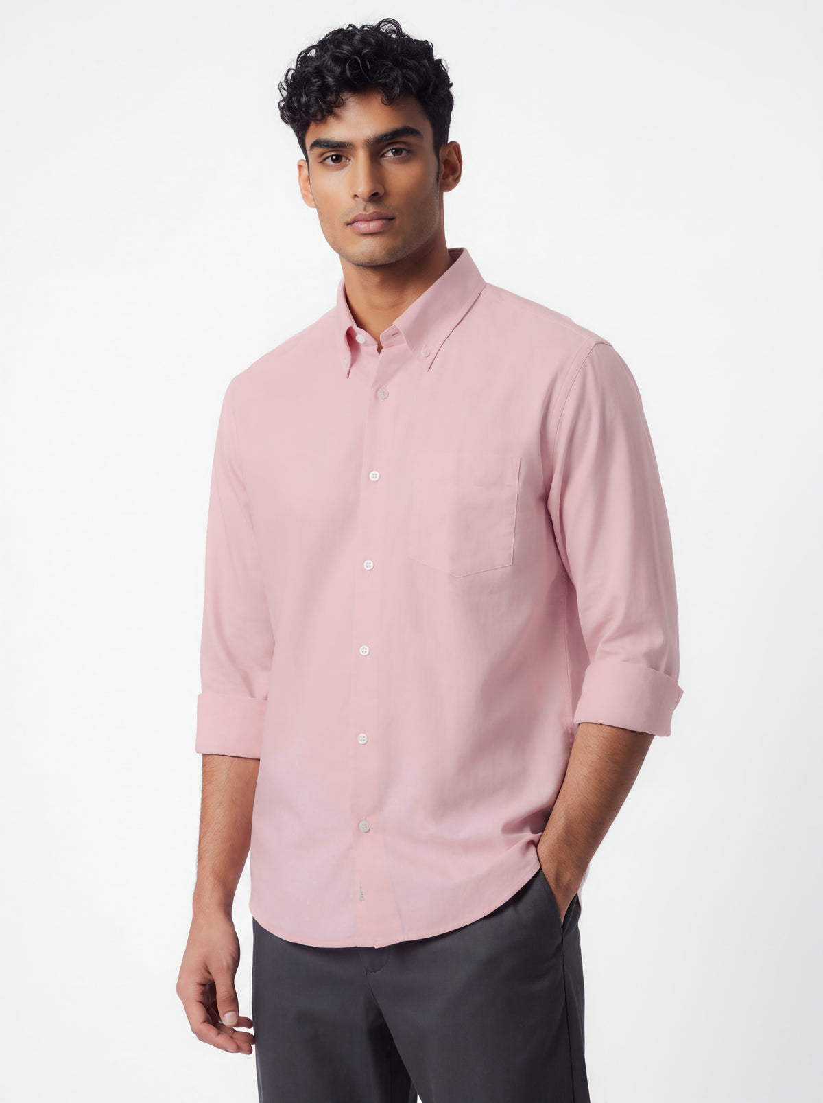 A man wearing a light pink long-sleeve button-down shirt with rolled-up sleeves and dark gray trousers, standing against a plain white studio background.