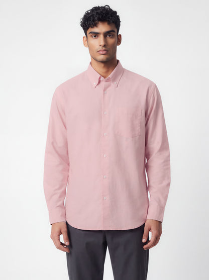 A man wearing a pastel pink  long-sleeve button-down shirt and dark gray trousers, standing against a plain light gray studio background.