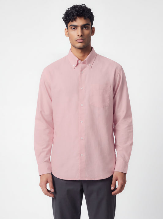 A man wearing a pastel pink  long-sleeve button-down shirt and dark gray trousers, standing against a plain light gray studio background.
