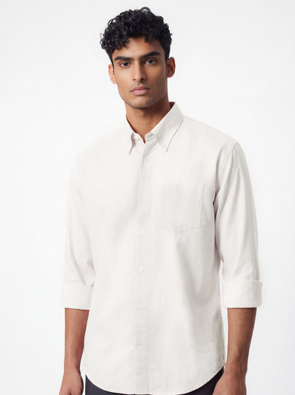 A man wearing a light beige long-sleeve button-down shirt and dark gray trousers, standing against a plain light gray studio background.