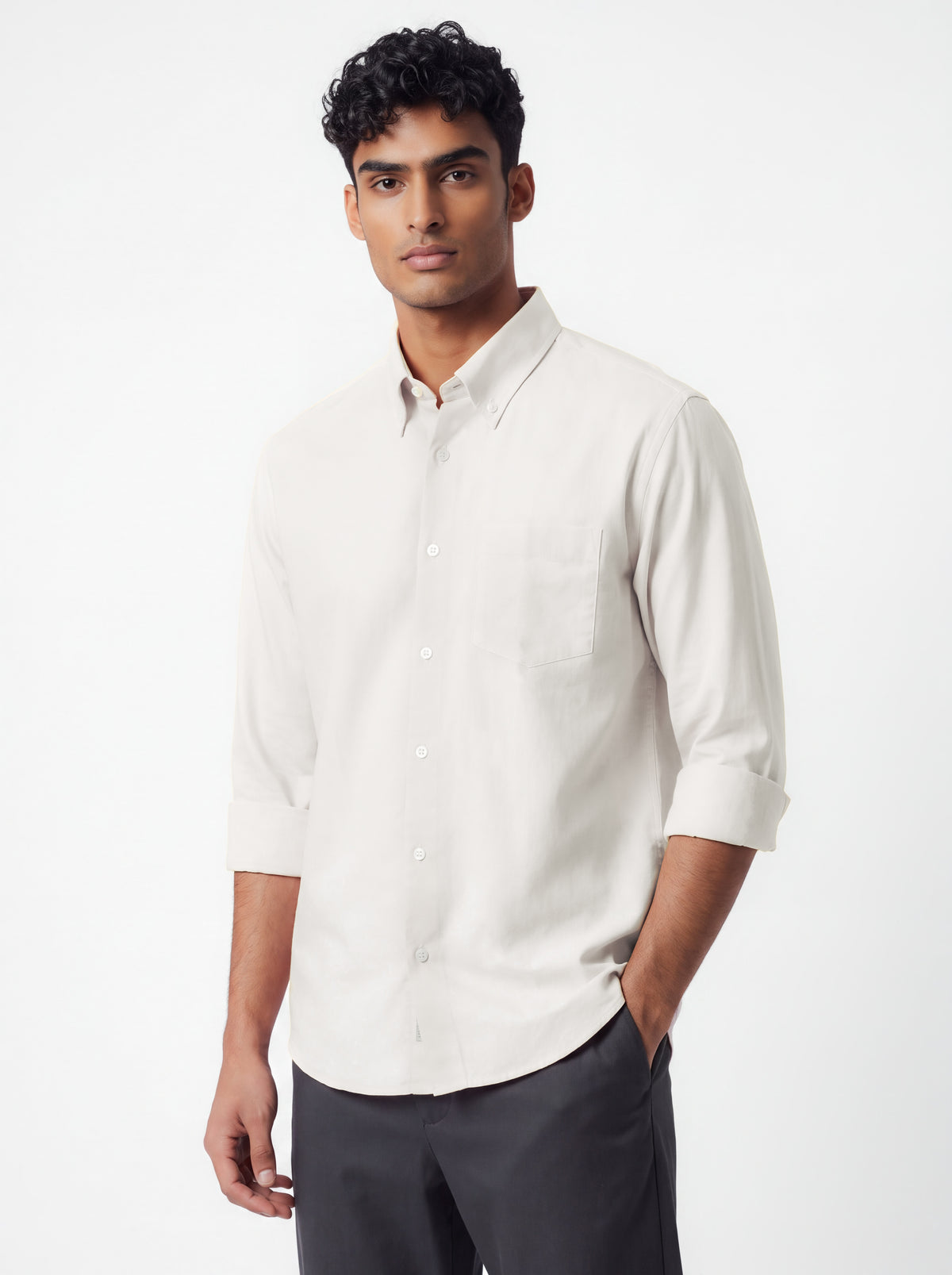 A man wearing a light beige long-sleeve button-down shirt and dark gray trousers, standing against a plain light gray studio background.