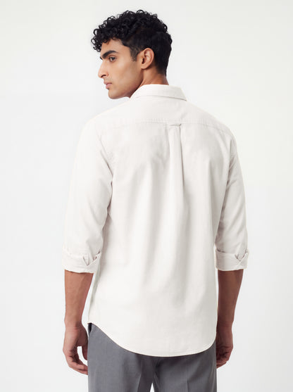 A man wearing a light beige long-sleeve button-down shirt and dark gray trousers, standing against a plain light gray studio background.