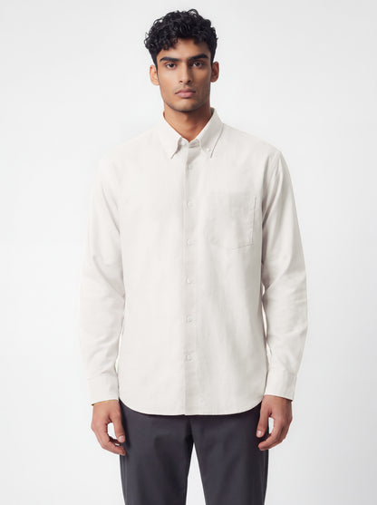 A man wearing a light beige long-sleeve button-down shirt and dark gray trousers, standing against a plain light gray studio background.