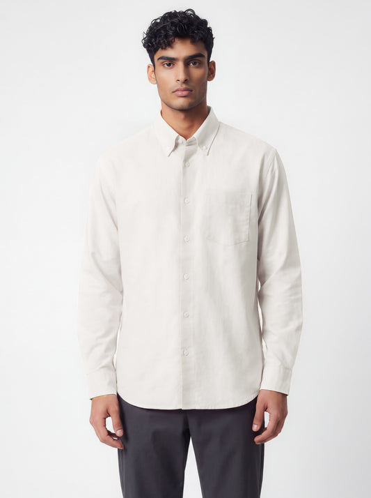 A man wearing a light beige long-sleeve button-down shirt and dark gray trousers, standing against a plain light gray studio background.