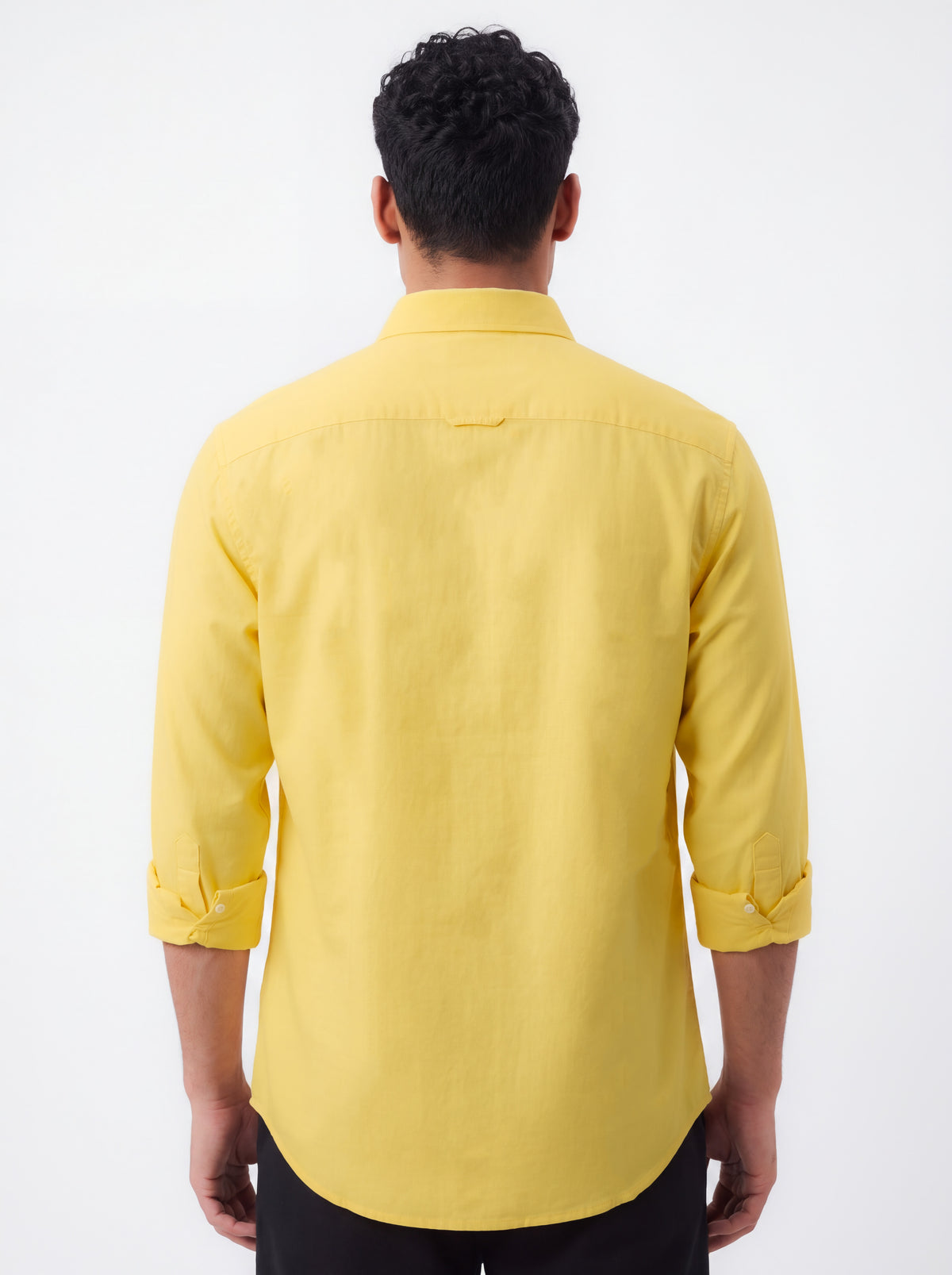 A man wearing a pastel yellow long-sleeve button-down shirt and dark gray trousers, standing against a plain light gray studio background.