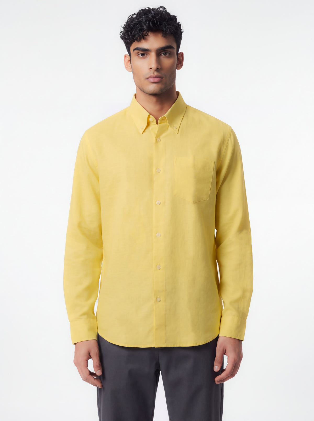 A man wearing a light yellow long-sleeve button-down shirt and dark gray trousers, standing against a plain white studio background.
