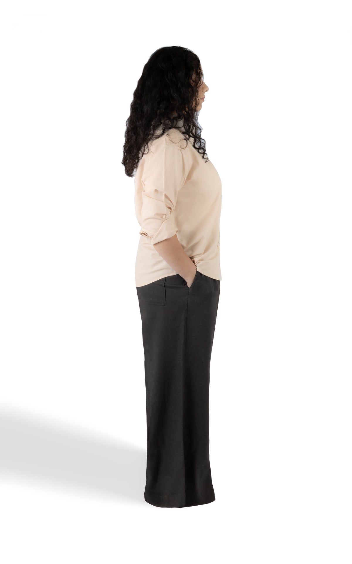A woman shown in side profile wearing a light beige long-sleeve top and dark charcoal gray wide-leg pants against a plain white studio background.