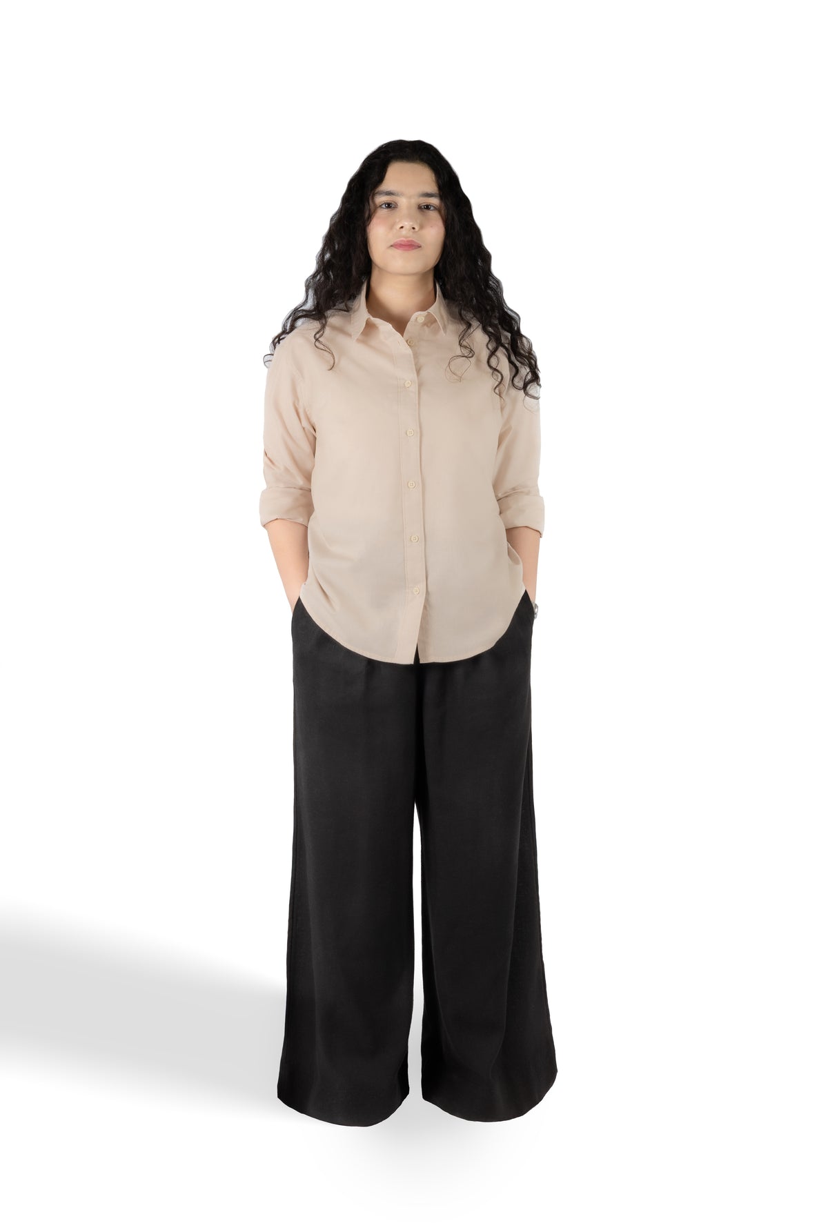 A woman stands against a white studio background wearing a light beige button-up shirt with rolled sleeves and black wide-leg pants.