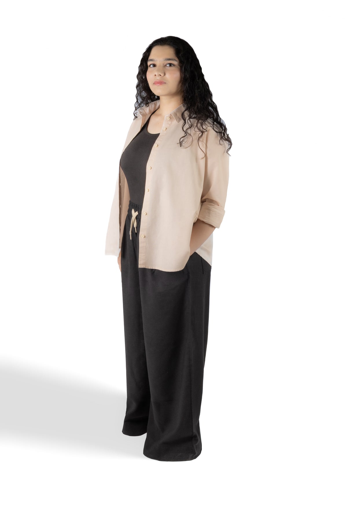 A woman modeling a light beige button-up shirt over a black top and black wide-leg pants, standing against a plain white studio background.