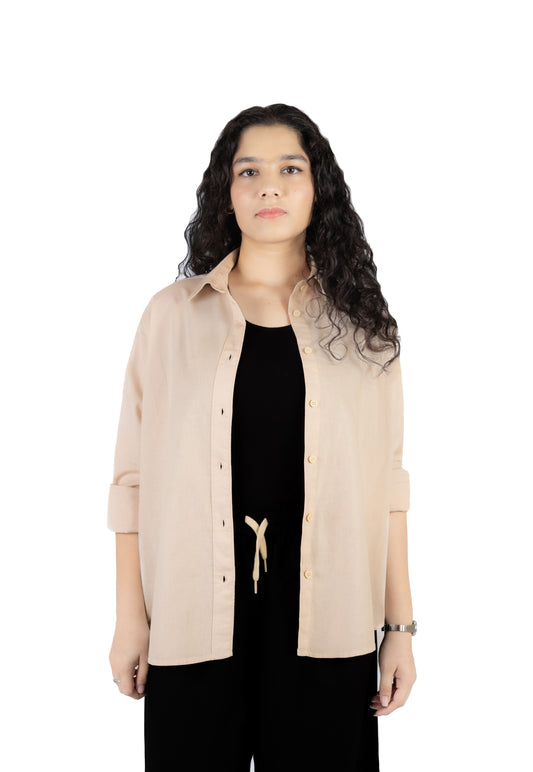 A woman with long, curly dark hair stands against a solid white background, wearing a light beige button-up shirt over a black top and black pants.