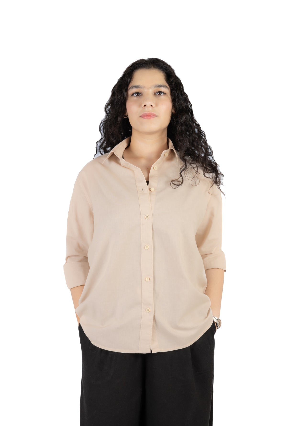 A woman with long, curly dark hair is wearing a light beige button-up shirt with rolled-up sleeves and black pants, standing against a solid white background.