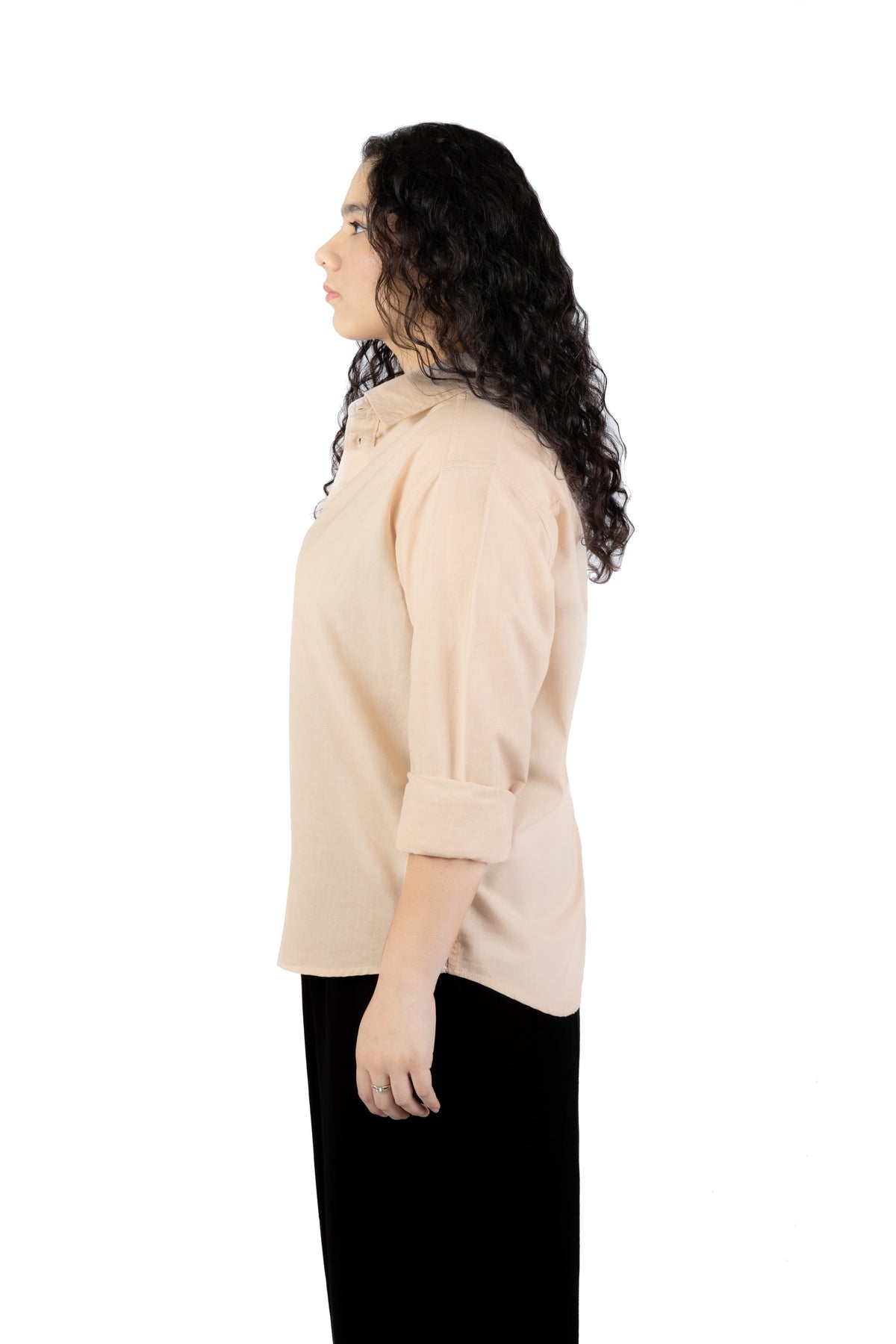 A woman with long, curly dark hair is shown in profile wearing a light beige or cream-colored blouse with rolled three-quarter sleeves and black pants, set against a solid white background.