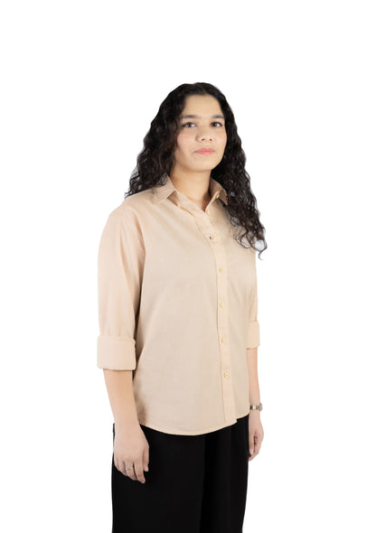 A woman with long, curly dark hair wearing a light beige button-up shirt with rolled-up sleeves and black pants, standing against a solid white background.
