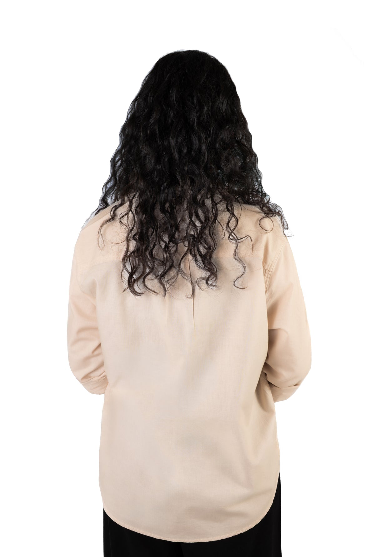 A person viewed from the back wearing a long, wavy black wig and a light beige long-sleeve top against a plain white background.