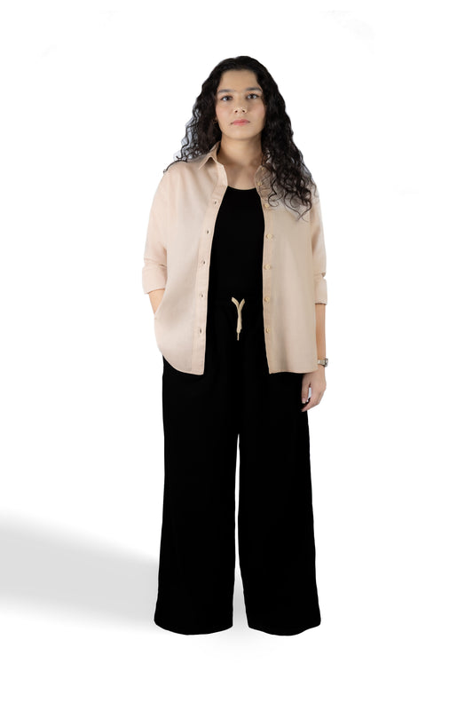 A woman modeling a light beige button-up shirt over a black jumpsuit, standing against a plain white studio background.