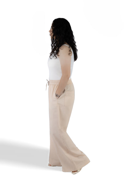 A woman shown from the side profile wearing a white sleeveless top and light beige wide-leg drawstring pants, standing against a plain white studio background.