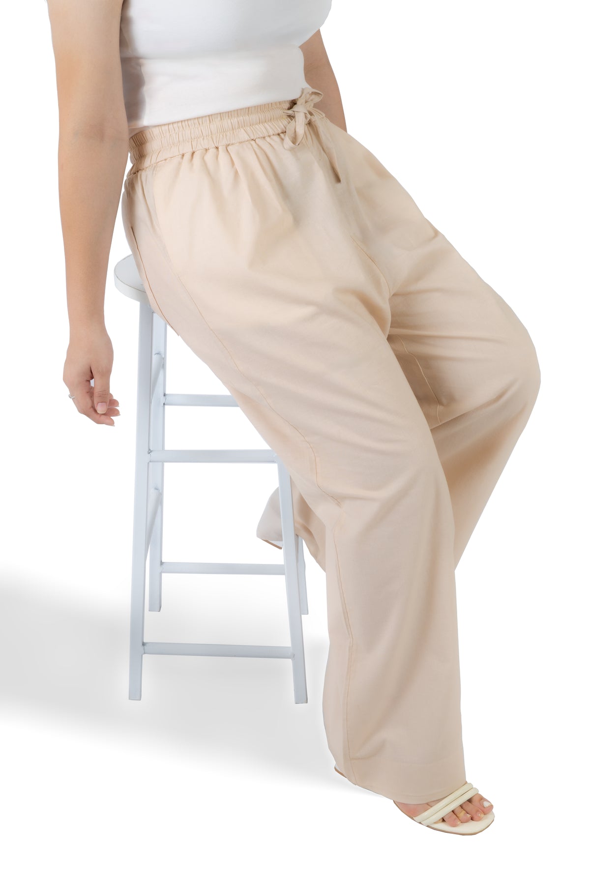 A person seated on a white stool wearing loose, wide-leg beige pants with an elastic drawstring waist, paired with a white top and light sandals, against a clean white background.