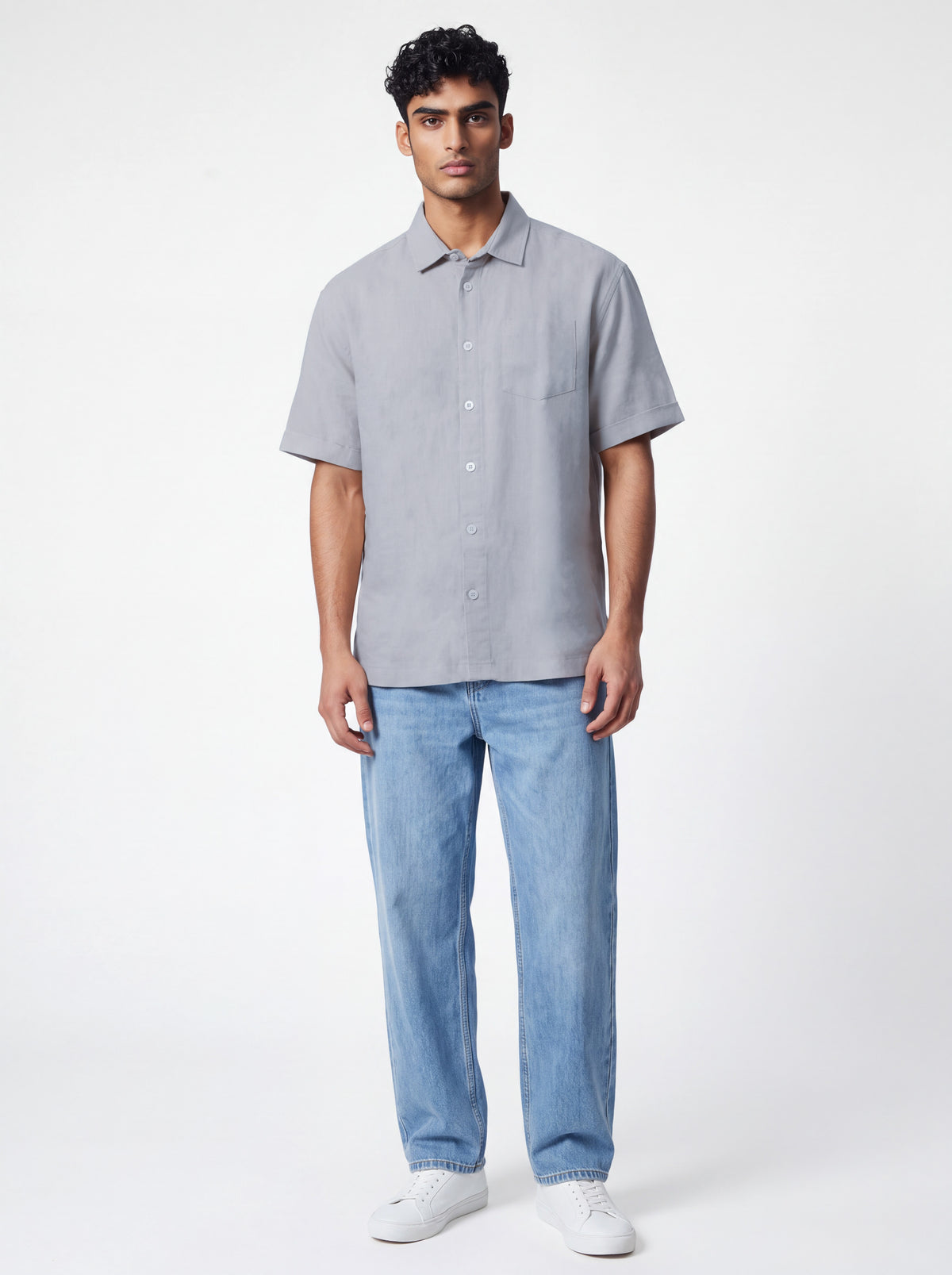 A man modeling a light gray short-sleeve button-up shirt with light blue jeans and white sneakers, standing against a plain white studio background.
