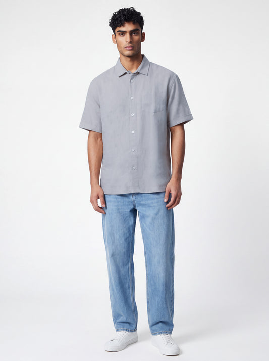 A man modeling a light gray short-sleeve button-up shirt with light blue jeans and white sneakers, standing against a plain white studio background.