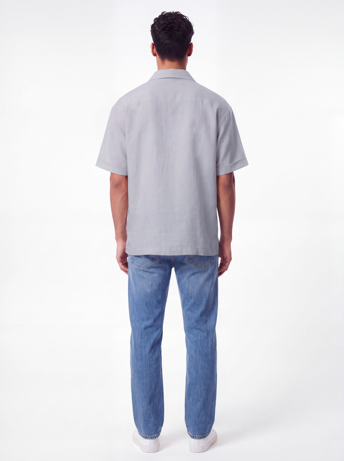 A man viewed from behind wearing a light gray short-sleeve button-up shirt and blue jeans against a plain white studio background.