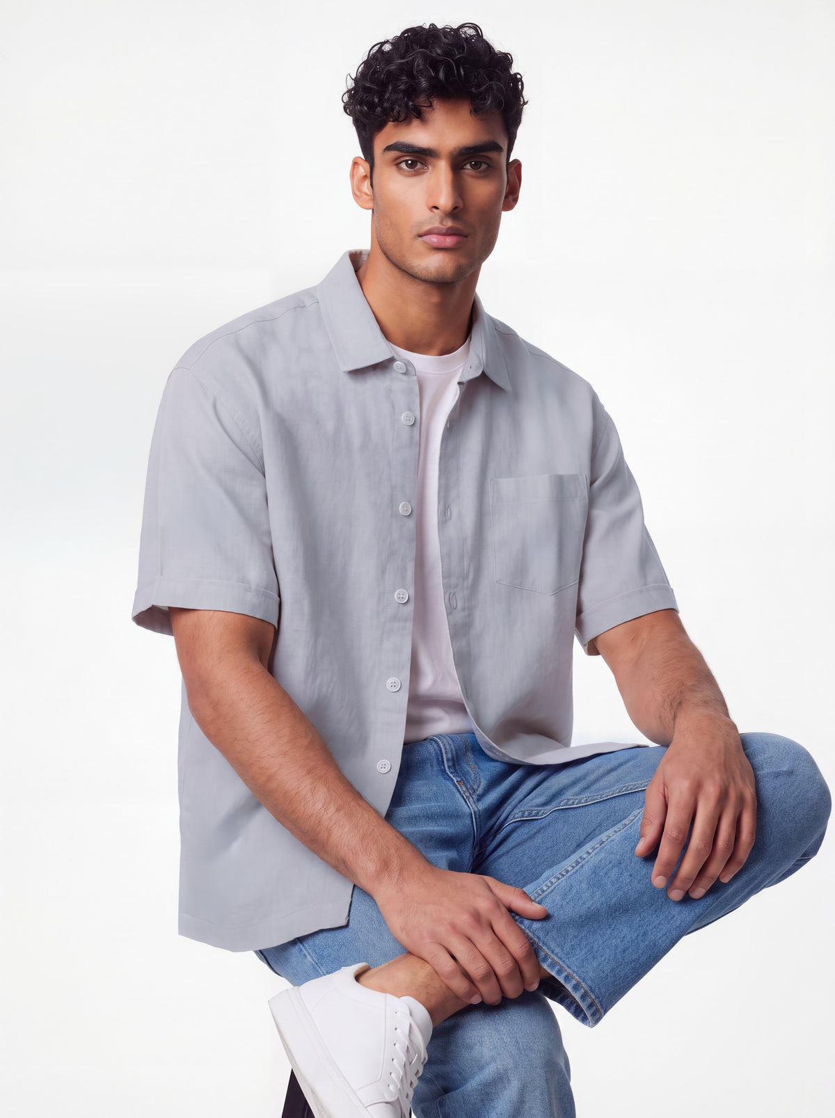 A man wearing a light gray short-sleeve button-up shirt over a white t-shirt, blue jeans, and white sneakers, posing against a plain white studio background.