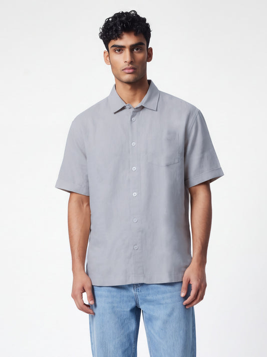 A man wearing a light gray short-sleeve button-up shirt and light blue jeans, standing against a plain white studio background.