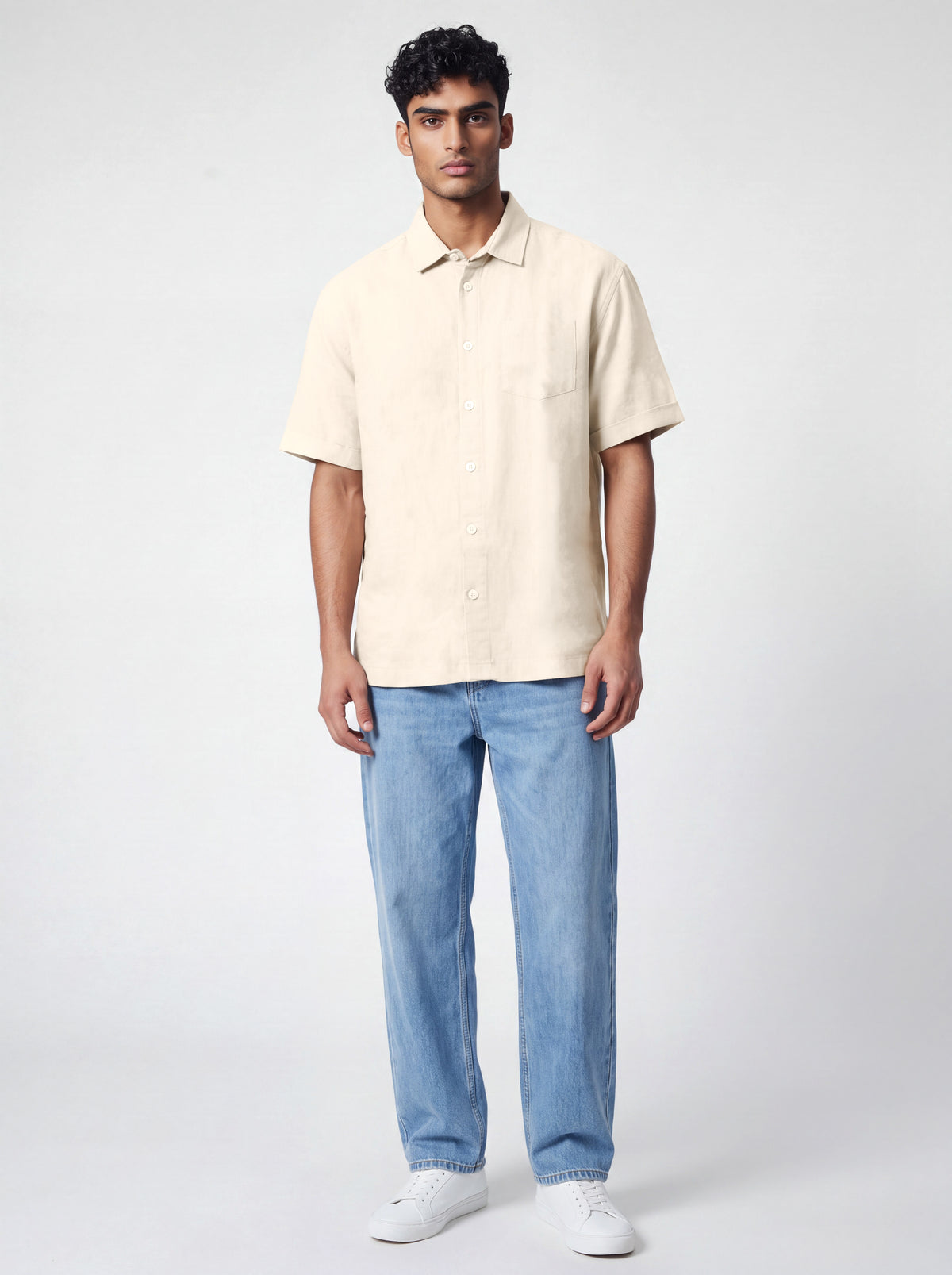 A man modeling a short-sleeve cream-colored linen shirt paired with light blue jeans and white sneakers, standing against a light gray studio background.