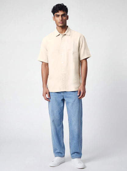 A man modeling a short-sleeve cream-colored linen shirt paired with light blue jeans and white sneakers, standing against a light gray studio background.