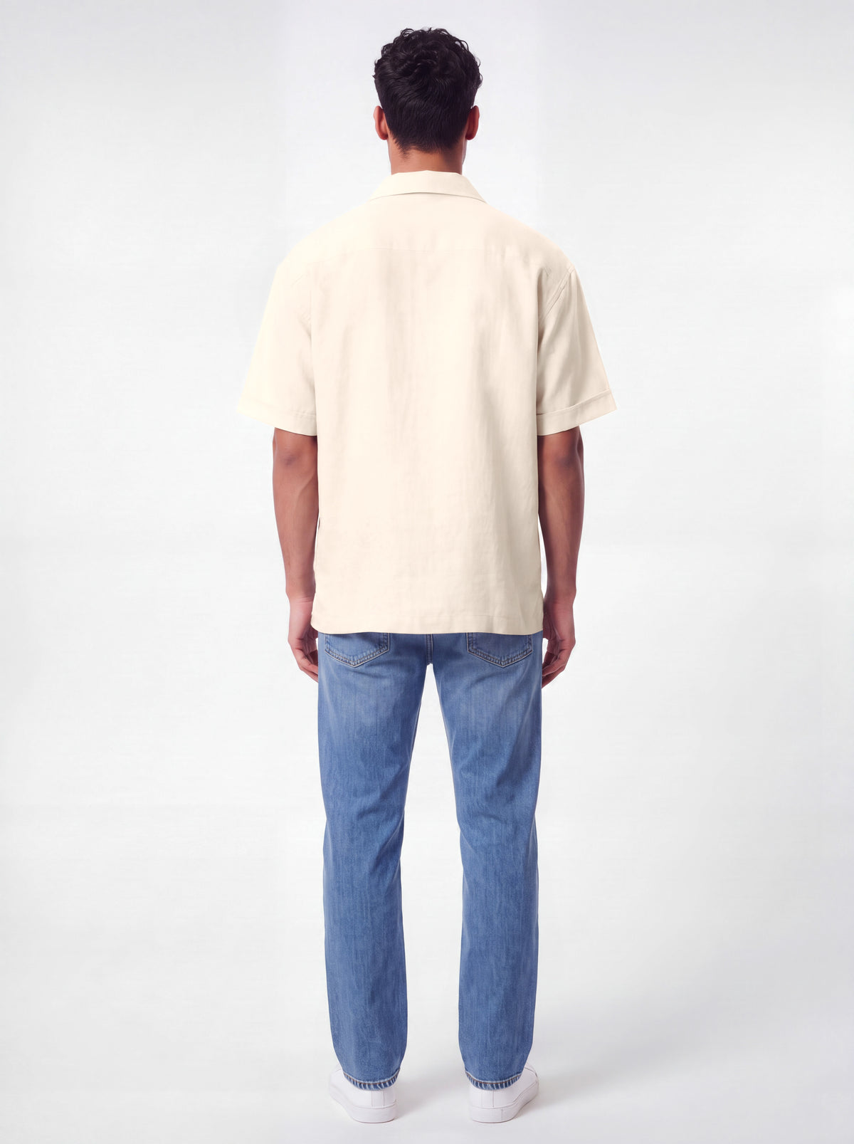 A man viewed from behind wearing a short-sleeve cream-colored linen shirt and blue jeans, standing against a plain white studio background.