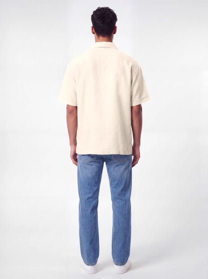 A man viewed from behind wearing a short-sleeve cream-colored linen shirt and blue jeans, standing against a plain white studio background.