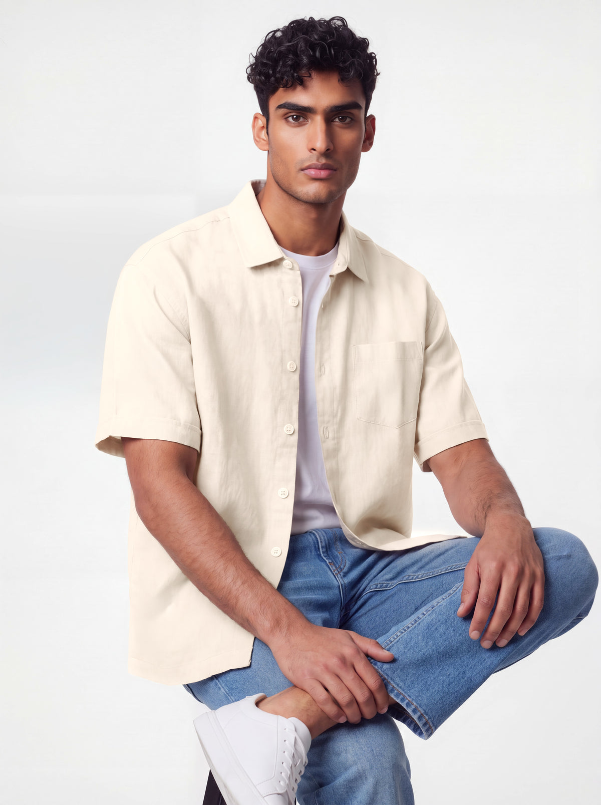 A man wearing a light beige short-sleeve linen button-up shirt over a white t-shirt, paired with blue jeans and white sneakers, posing against a light gray studio background.